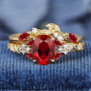 May include: A gold ring set featuring a central oval-cut red gemstone surrounded by smaller marquise-cut red stones and round white stones. The band is designed with a sculpted leaf and vine motif, incorporating additional small white stones.
