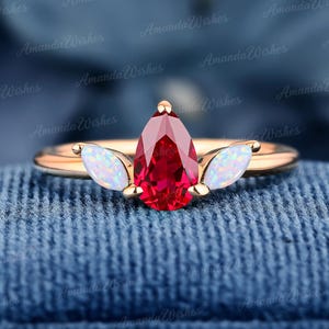 May include: A rose gold ring featuring a central pear-cut red gemstone flanked by two marquise-cut iridescent white opal stones. The band is smooth and polished, showcasing a three-stone design with a vibrant red centre and light-reflecting side accents.