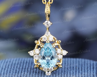 May include: A gold pendant necklace featuring a central pear-shaped light blue gemstone surrounded by small round clear stones in a decorative setting. The pendant hangs from a delicate gold chain, with a small clear stone accent above the main piece.
