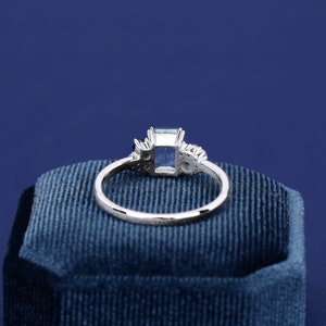 May include: A silver ring with a light blue square-shaped gemstone surrounded by smaller clear gemstones. The ring is sitting on a blue velvet box.