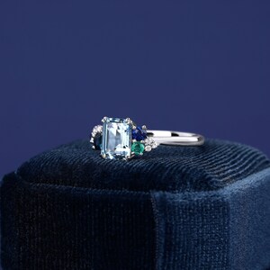 May include: A silver ring with a large emerald-cut aquamarine gemstone surrounded by smaller blue and green gemstones. The ring is sitting on a blue velvet box.