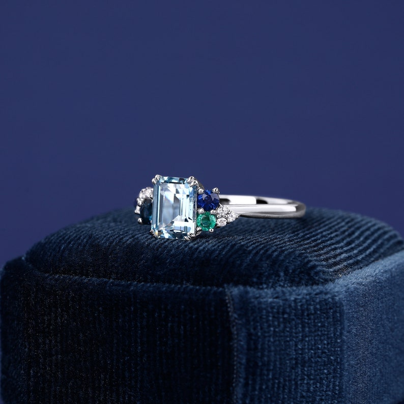 May include: A silver ring with a large emerald-cut aquamarine gemstone surrounded by smaller blue and green gemstones. The ring is sitting on a blue velvet box.