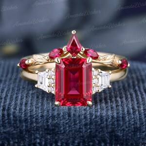 May include: A gold ring set featuring a central emerald-cut red gemstone flanked by two baguette-cut white stones and small round accents. A matching curved band sits above, adorned with marquise-cut red stones and gold leaf detailing.
