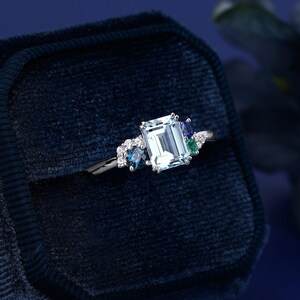May include: A silver ring with an emerald-cut aquamarine center stone surrounded by smaller blue and green gemstones. The ring is displayed in a blue velvet ring box.