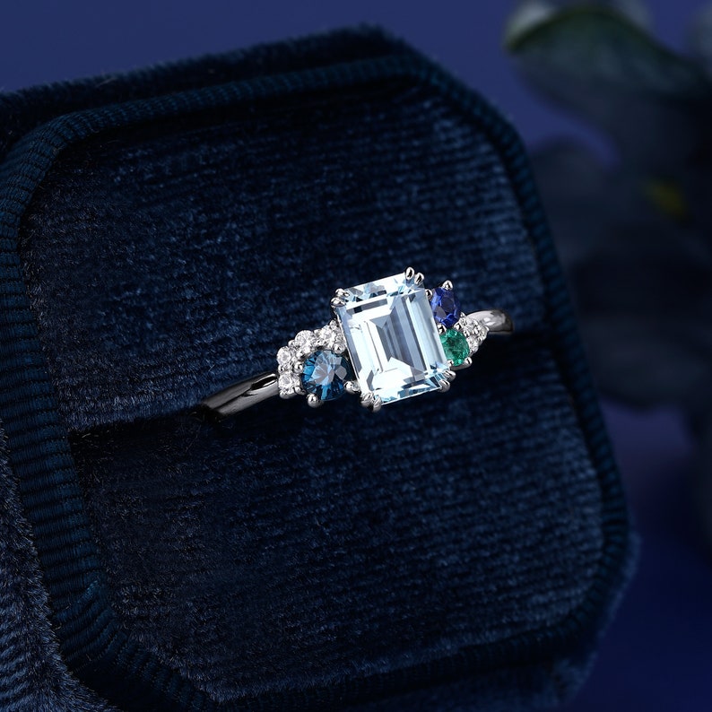 May include: A silver ring with an emerald-cut aquamarine center stone surrounded by smaller blue and green gemstones. The ring is displayed in a blue velvet ring box.