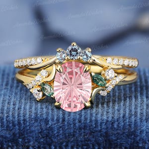 Puede incluir: Un juego de anillos de compromiso de oro con una gran piedra rosa de corte ovalado, acentuada con pequeñas piedras azules y verdes. La banda está adornada con pequeñas piedras transparentes. El texto "Amanda Wishes" es visible.
