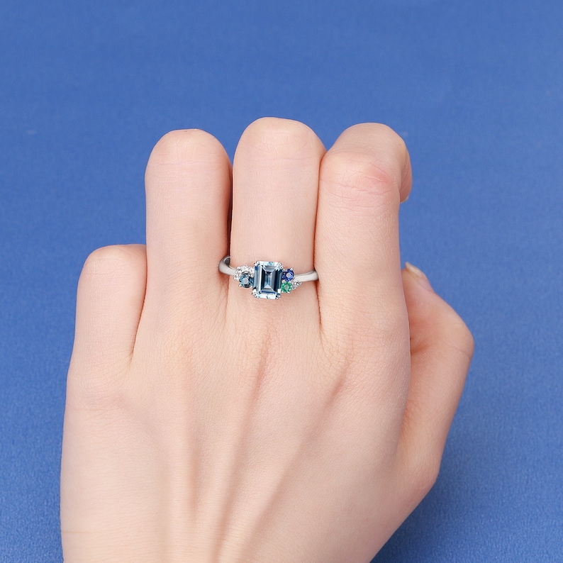 May include: A silver ring with a light blue emerald-cut gemstone surrounded by smaller blue and green gemstones.