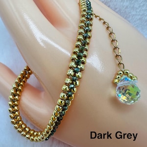 May include: A gold and dark grey beaded bracelet with a crystal charm hanging from a chain. The bracelet is on a hand, and the text "Dark Grey" is visible below the bracelet.