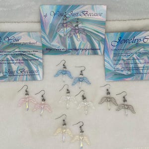 Puede incluir: Ocho pares de pendientes de alas de &aacute;ngel en varios colores, incluyendo azul, blanco y rosa. Los pendientes se muestran sobre una superficie blanca con un fondo hologr&aacute;fico. El texto "You Just Because" es visible en las tarjetas detr&aacute;s de los pendientes.