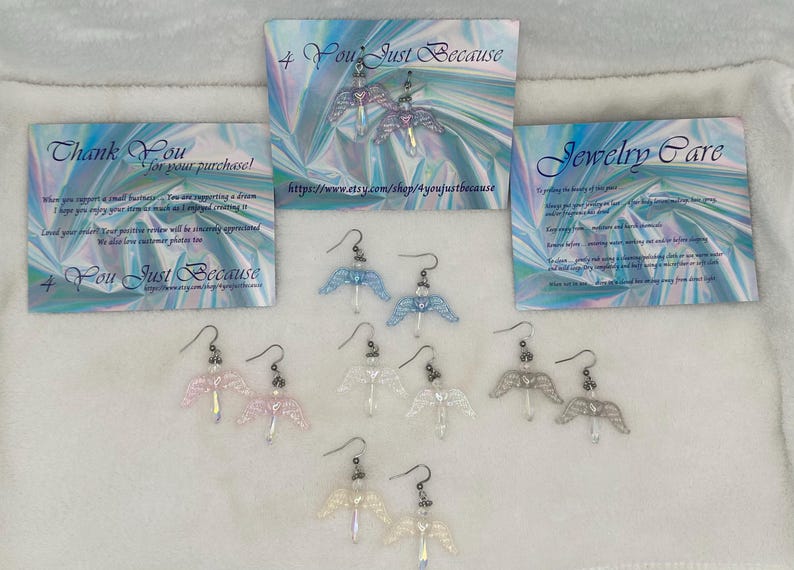 Puede incluir: Ocho pares de pendientes de alas de &aacute;ngel en varios colores, incluyendo azul, blanco y rosa. Los pendientes se muestran sobre una superficie blanca con un fondo hologr&aacute;fico. El texto "You Just Because" es visible en las tarjetas detr&aacute;s de los pendientes.