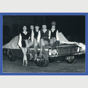 May include: Five women wearing hats and dresses are sitting on the bonnet of a black car. The car has a white tarp covering the windscreen and bonnet. The car has a number plate that reads "KHB-375".
