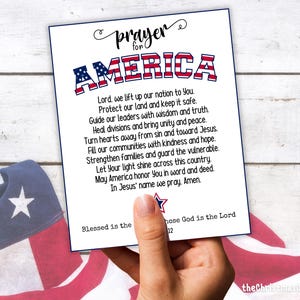 May include: A white card with the words "prayer for AMERICA" in red, white, and blue. The card contains a prayer for the United States. The card is held in a hand, with an American flag in the background.