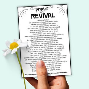 May include: A white card with the text "prayer for REVIVAL" is held by a hand, with a daisy in the foreground. The card contains a prayer. The background is a light blue colour.