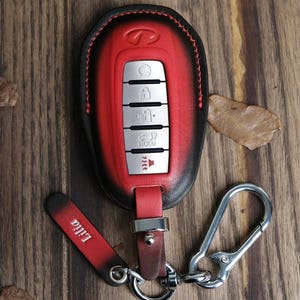 May include: Red and black leather car key fob cover with silver buttons and a keychain. The fob has the Infiniti logo and a leather tag that says "Lilia". The key fob is on a wooden surface with leaves.