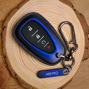 May include: A blue and black leather car key fob cover featuring a metallic carabiner clip and a small blue leather tag with the word Carder embossed in white. The key fob has four buttons with icons for locking, unlocking, and remote engine start.