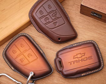 May include: Three brown leather key fob covers with visible stitching. One cover features the Chevrolet Tahoe logo embossed on the surface, while the other two show button icons. A silver metal carabiner is attached to one of the covers.