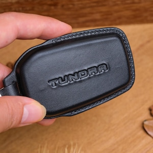 Puede incluir: Llavero de cuero negro con la palabra "TUNDRA" en relieve. El llavero es rectangular con esquinas redondeadas y un borde cosido. Un anillo de metal plateado está unido al llavero.
