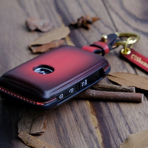 May include: A red leather key fob with a stitched edge and a silver key. The fob has a small silver button and a silver keyhole. The fob is attached to a gold key ring with a small silver charm.