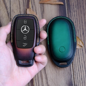 May include: Two leather key covers, one brown with a Mercedes-Benz logo and the other green. The covers are stitched with a contrasting thread.