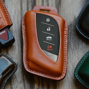Puede incluir: Un llavero de cuero marrón con costuras alrededor de los bordes. El llavero tiene un inserto negro con botones etiquetados como "Hold" y un icono de coche. El llavero está sobre una superficie de madera.