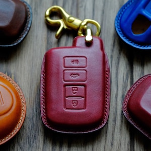 May include: A close-up of a red leather key fob with three buttons. The fob is attached to a gold-coloured key ring. The key fob is surrounded by other leather key fobs in different colours.