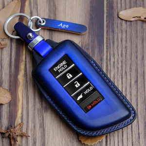 May include: A blue leather key fob with a silver key ring and a name tag that says "Aga". The fob has buttons that say "Engine Hold" and "Hold".