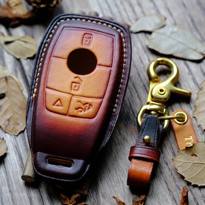 May include: A brown leather key fob with a gold metal keychain and a leather strap. The key fob has a cutout for a car key and four buttons. The keychain has a gold metal clasp and a leather tag with the letter "T" on it.