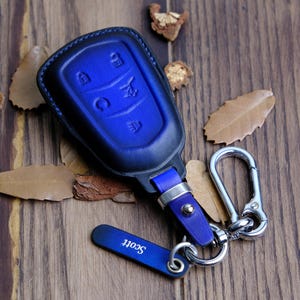Puede incluir: Llavero de cuero azul con un anillo y un gancho plateados. El llavero tiene cuatro botones y el texto "Scott" está grabado en el gancho.