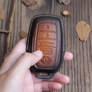 May include: A brown and black leather car key fob cover with a Toyota logo. The cover has embossed buttons for lock, unlock, and other functions. The key fob is held in a hand.