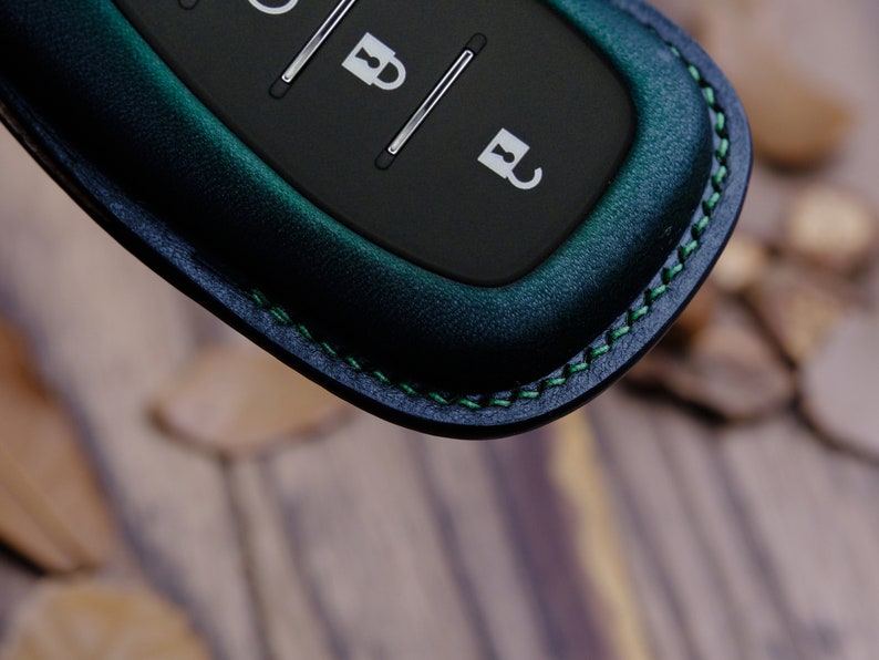 May include: A dark green leather key fob with a black remote control. The fob has green stitching detail around the edge.