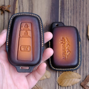 Könnte beinhalten: Zwei braune Leder-Schlüsselhüllen. Eine hat ein Schlüsselanhänger-Design und die andere hat den Text "4Runner" geprägt.