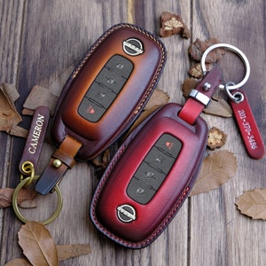 May include: Two leather key fobs with a red and brown color gradient. The key fobs have a Nissan logo and four buttons. One key fob has a gold keychain with a tag that says "CAMERON". The other key fob has a gold keychain with a tag that says "201-370-3486".