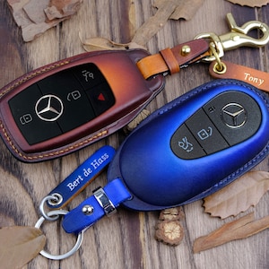 May include: Two leather keychains with key fobs. One keychain is brown leather with a silver Mercedes-Benz logo and the text "Bert de Hass". The other keychain is blue leather with a silver Mercedes-Benz logo. Both keychains have a silver ring.