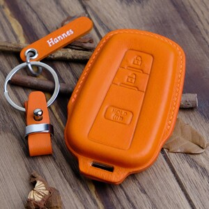 Könnte beinhalten: Orangefarbener Schlüsselanhänger aus Leder mit einem silbernen Schlüsselring und einem Lederanhänger mit der Aufschrift "Hannes".