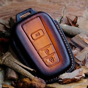 May include: A brown leather key fob with a stitched edge and three buttons. The buttons are embossed with symbols.