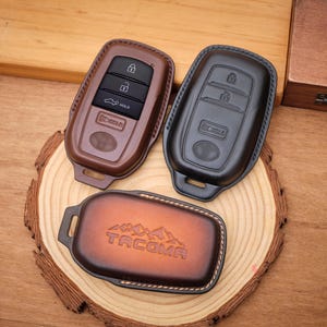May include: Three key fob covers for a vehicle. One cover is brown, one is black, and the third is a gradient brown with a mountain graphic and the word TACOMA embossed on the front. Each cover features stitched edges and a cutout for a key ring.