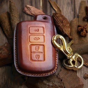 May include: A brown leather key fob with four buttons and a gold metal key chain. The key fob is sitting on a wooden surface with various pieces of wood and spices.