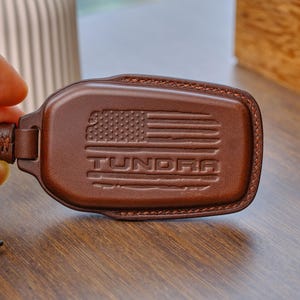 May include: A dark brown leather key fob cover featuring an embossed American flag design and the word TUNDRA centered below the flag. The cover is secured with a small leather strap and a gold-toned metal screw fastener on the left side.