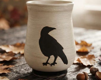May include: A cylindrical ceramic tumbler with a matte off-white finish and a slightly flared rim. The front features a solid black silhouette of a crow standing in profile. The base of the vessel is finished in a contrasting dark black glaze.