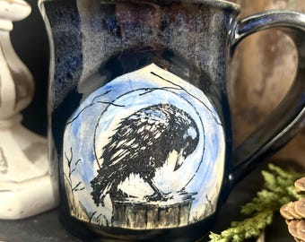 May include: A ceramic mug with a deep navy blue glaze featuring a hand-painted illustration of a black raven perched on a stump against a pale blue circular moon background. The mug has a curved handle and a smooth, glossy finish throughout.