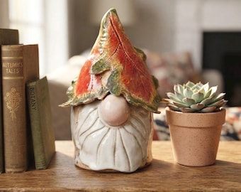 May include: A ceramic gnome figurine featuring a hat shaped like a large, textured autumn leaf in shades of orange, red, and green. The gnome has a round, pale pink nose and a long, white, textured beard. It sits beside a small succulent plant.