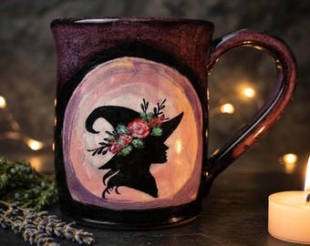 May include: A dark purple ceramic mug featuring a central circular illustration of a black silhouette wearing a pointed hat adorned with red and green floral accents. The mug has a glossy finish and a curved handle on the right side.