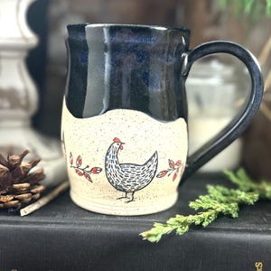 May include: A ceramic mug featuring a two-tone design with a glossy dark blue upper section and a speckled cream lower section. The cream portion is decorated with a hand-drawn illustration of a chicken and small red leaf sprigs.
