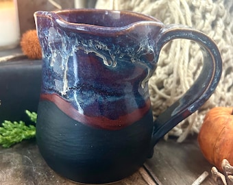 May include: A ceramic pitcher featuring a matte black base and a glossy upper section with a drip glaze effect in shades of deep blue, purple, and white. The vessel has a curved handle on the right side and a distinct pouring spout at the rim.