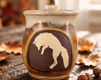 May include: A ceramic jar with a lid featuring a cream and brown speckled glaze. The front displays a dark brown circular inset with a light-colored silhouette of a fox in a leaping pose. The lid has a small, pointed, triangular handle.