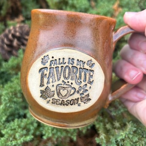 May include: A glossy, amber-brown ceramic mug with a rounded body and a side handle. A circular, light-colored inset on the front features the embossed text "FALL IS MY FAVORITE SEASON" surrounding a small graphic of a mug with a heart.
