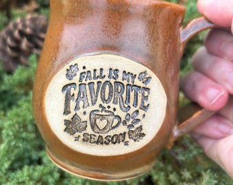 May include: A glossy, amber-brown ceramic mug with a rounded body and a side handle. A circular, light-coloured inset on the front features the embossed text "FALL IS MY FAVORITE SEASON" surrounding a small graphic of a mug with a heart.