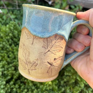 Puede incluir: Taza de cerámica hecha a mano con borde y asa esmaltados en azul claro. La parte inferior de la taza es de color beige con diseños de hojas en relieve. Una taza de café o té rústica y única.