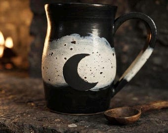 May include: A black ceramic mug featuring a white, speckled cloud-like design on the front with a solid black crescent moon shape in the center. The mug has a curved handle and a smooth finish, accompanied by a small, dark wooden spoon.