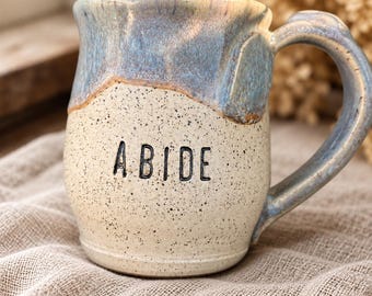 May include: A ceramic mug with a speckled cream-colored base and a glossy blue glazed rim. The word ABIDE is stamped in black capital letters on the front. The mug features a curved handle and a rounded body with a textured, hand-crafted appearance.